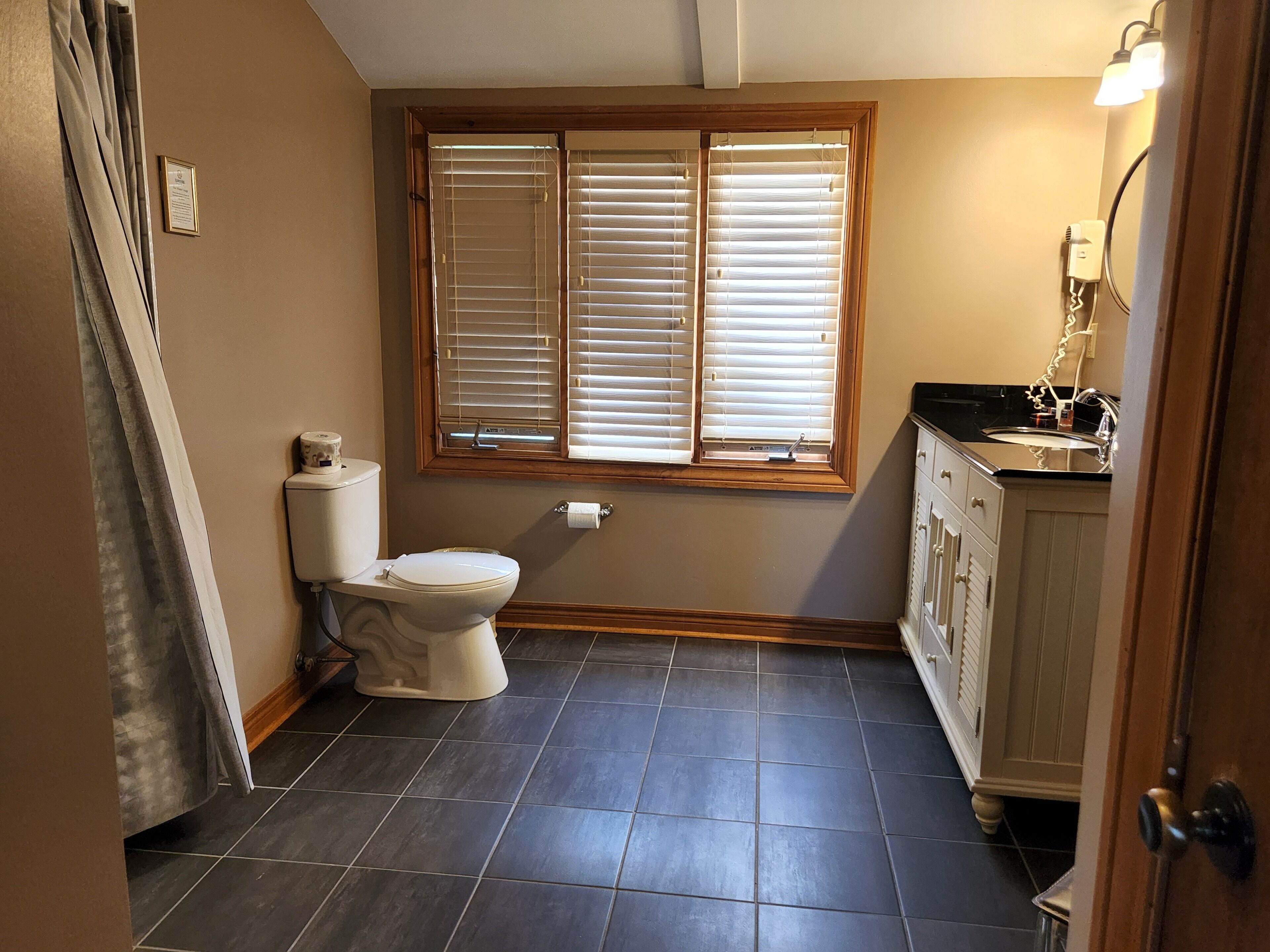 A bathroom with a toilet, a window with blinds, a sink, and a cabinet.