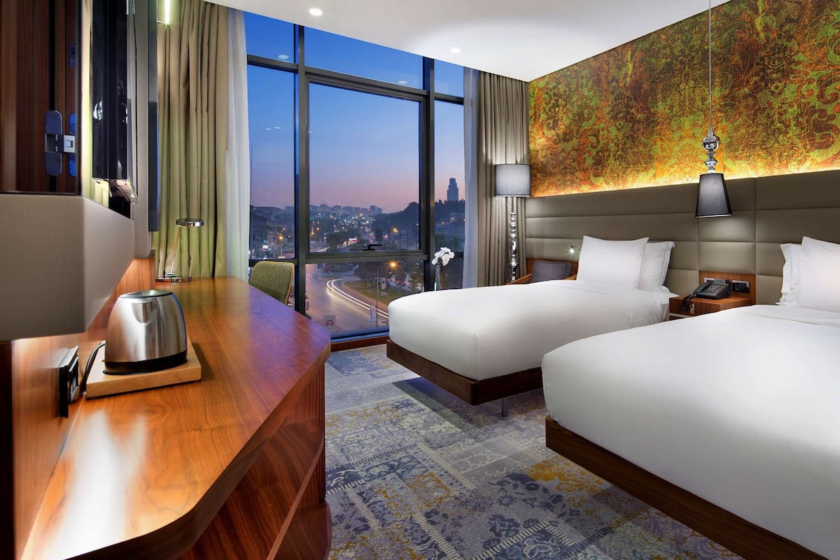 A hotel room with two beds, a wooden desk with a kettle, a nightstand, and a large window offering a city view.