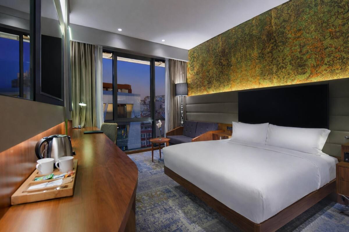 A hotel room with a large bed, a desk with a kettle and cups, a sofa, and a view of the city through large windows.