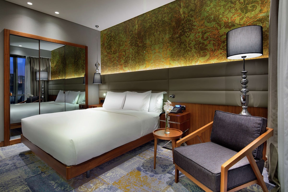 A modern hotel room with a large bed, a sitting area with a chair, a small table, and a mirror.