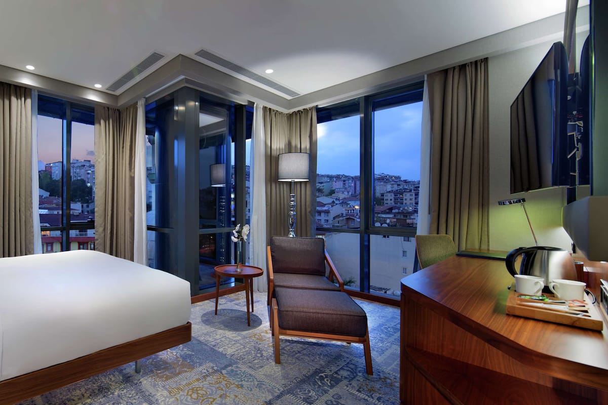 A hotel room with a large bed, a sitting area with a chair and a table, a desk with a lamp, and a view of the city through large windows.