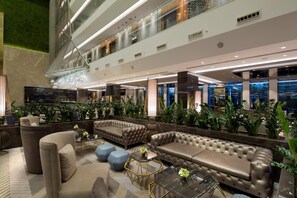 Eine geräumige Lobby mit hoher Decke, mehreren Sitzbereichen und Blick auf die Stadt durch große Fenster.