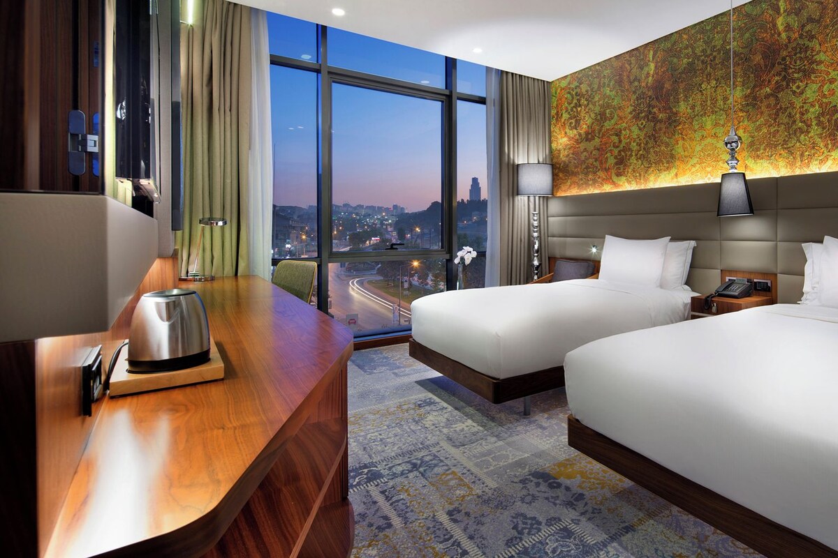 A hotel room with two beds, a desk with a kettle, a telephone, and a city view.