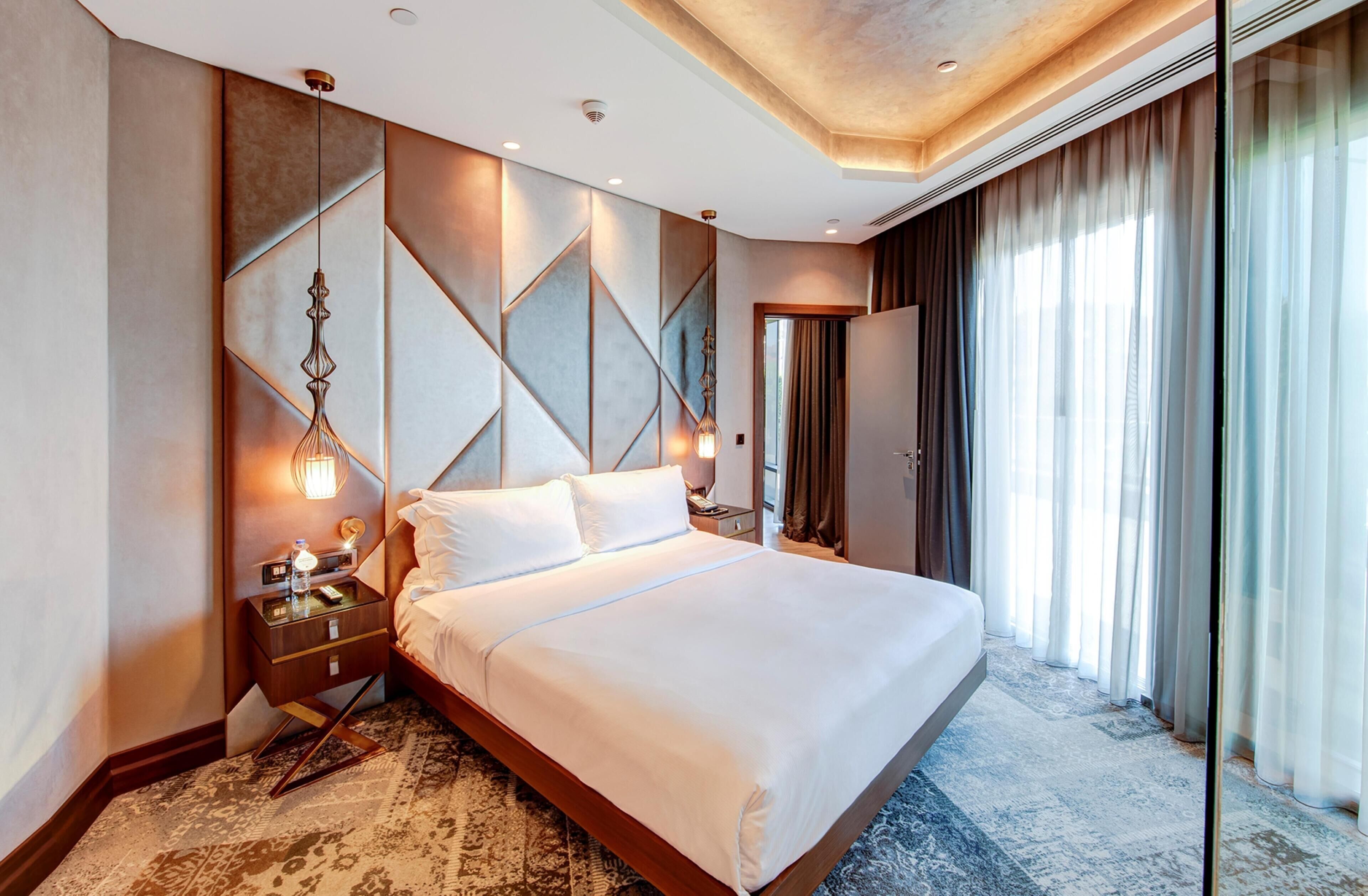 A modern hotel room with a large bed, a nightstand, a decorative wall, and a view of the outdoors.