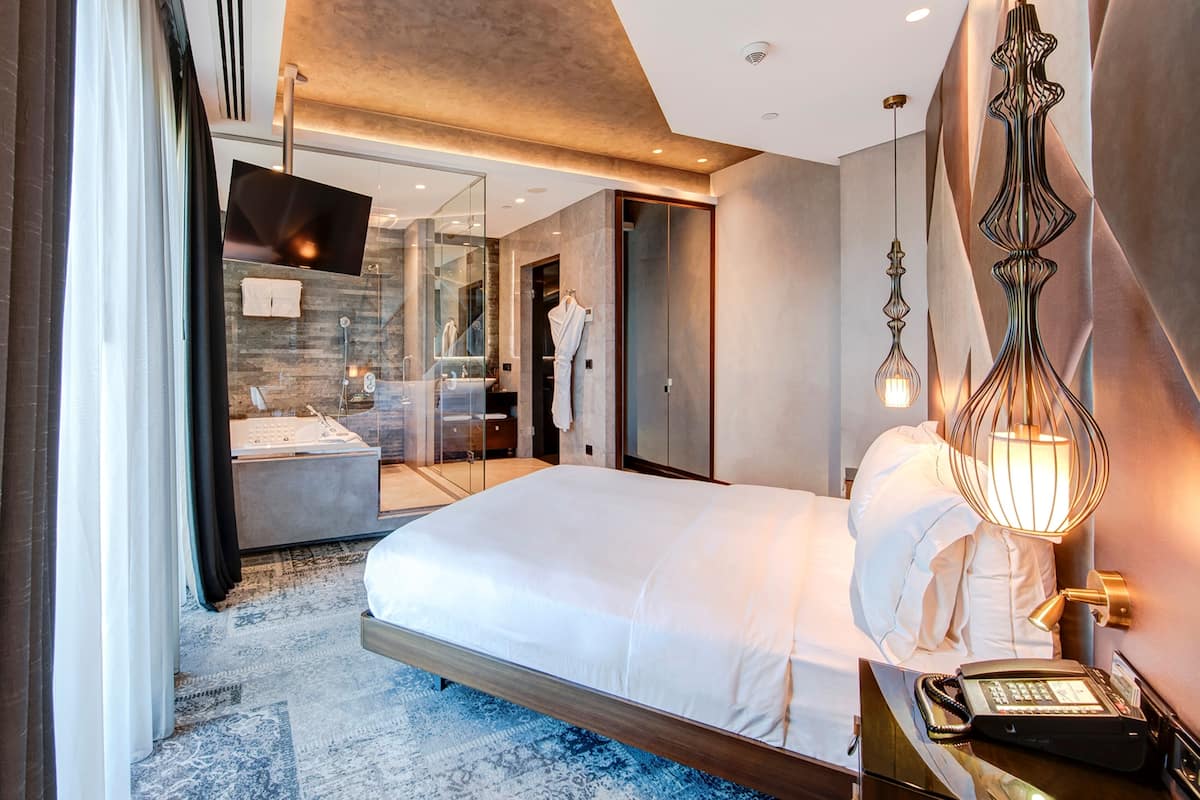 A modern hotel room with a large bed, a glass-enclosed shower, a vanity with a mirror, and a hanging light fixture.