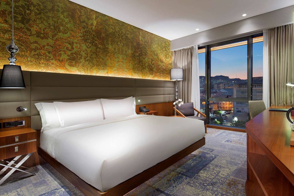 A hotel room with a large bed, a desk with a coffee maker, a chair, and a view of the cityscape through large windows.