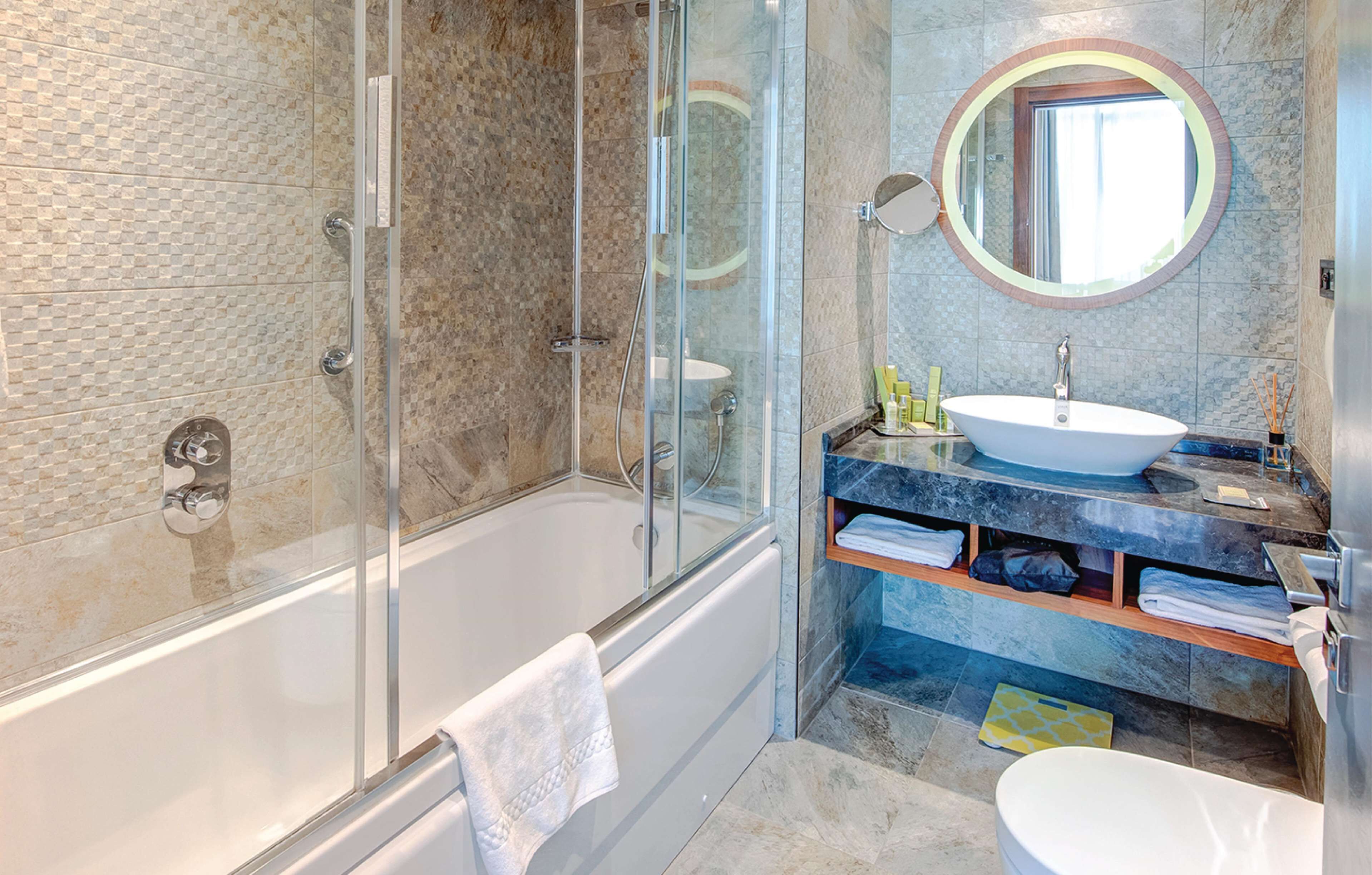 A modern bathroom with a glass-enclosed shower, a freestanding bathtub, a round mirror, and a marble vanity with a sink.