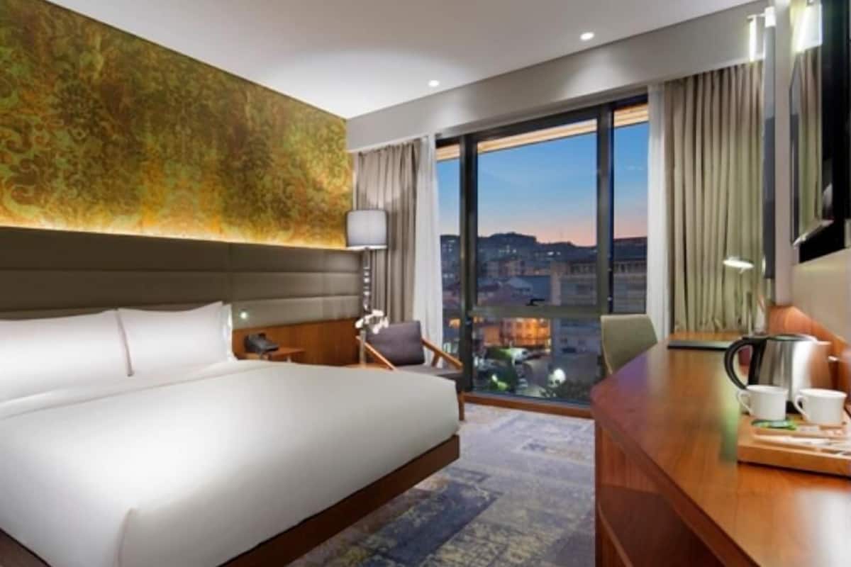 A hotel room with a large bed, a desk with a coffee maker, a chair, a nightstand, and a view of the cityscape through large windows.