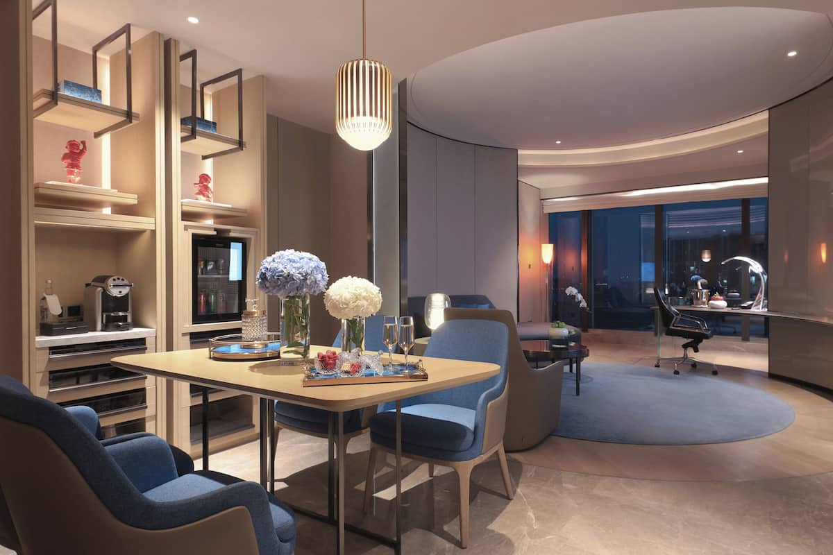 A modern hotel room with a dining area, a minibar, a TV, and a large window offering a view of the city.
