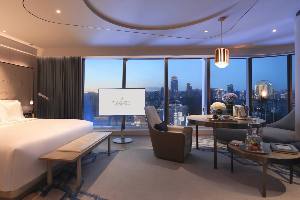 A modern hotel room with a large bed, a desk, and a view of the cityscape.