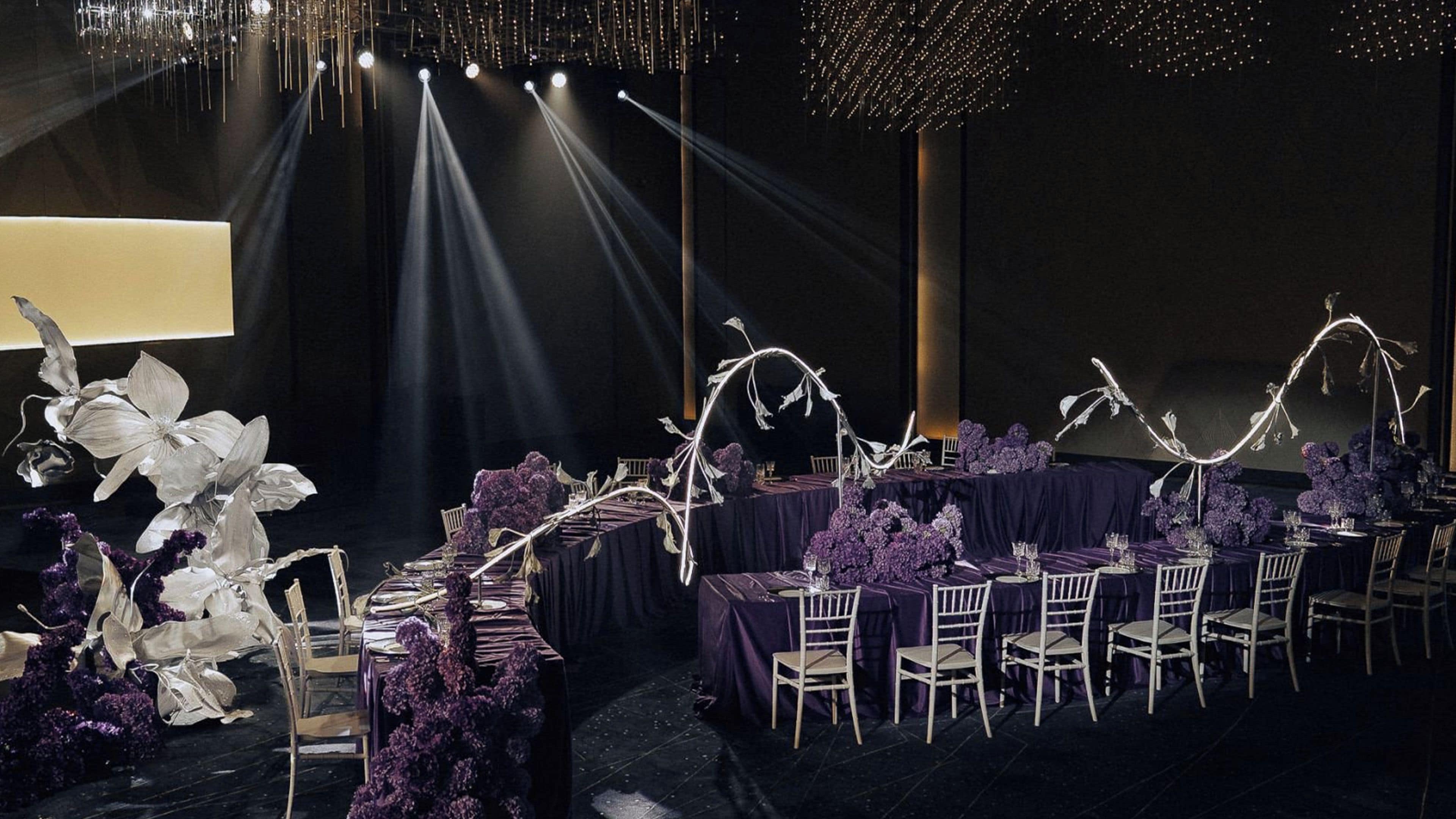 A formal dining setup with tables covered in dark cloths, white chairs, and purple floral arrangements.
