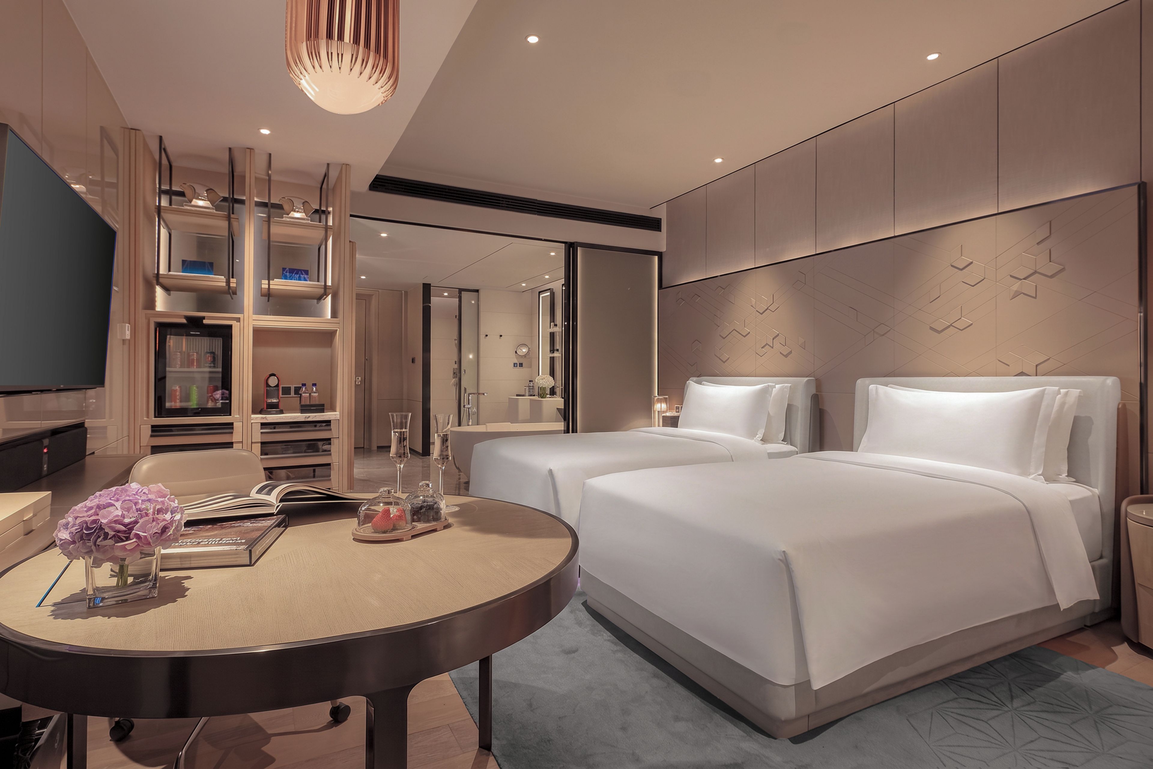 Une chambre d’hôtel moderne comprenant un lit, des tables de chevet, une table ronde avec un vase et un livre, un téléviseur et un dressing.