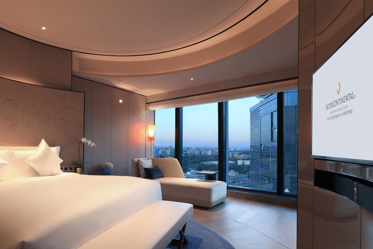 A modern hotel room with a large bed, a seating area, a city view, and a large screen displaying the hotel name.