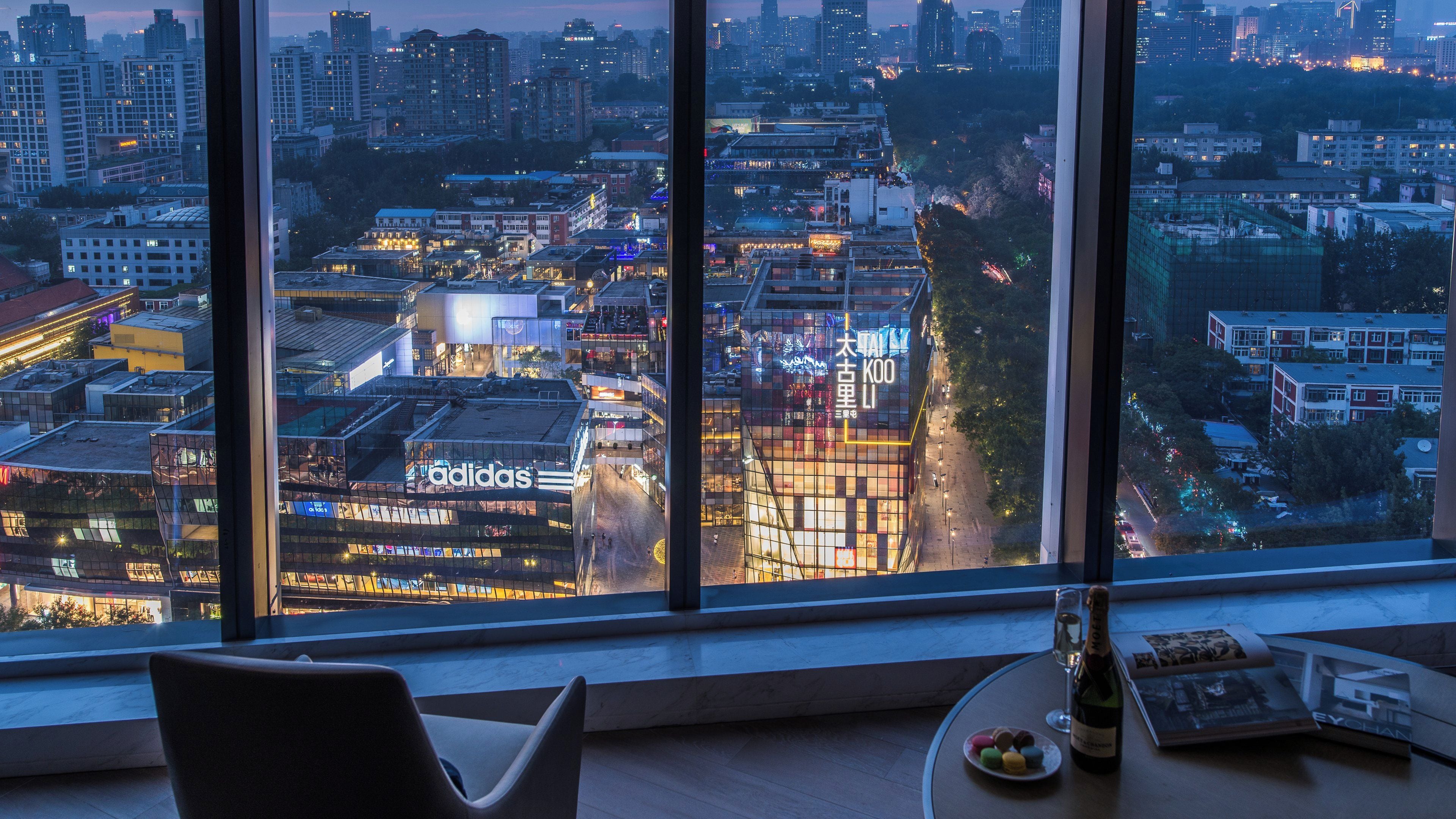 Une chambre d’hôtel située dans un immeuble de grande hauteur, avec une grande fenêtre donnant sur une vue panoramique de la ville au crépuscule.