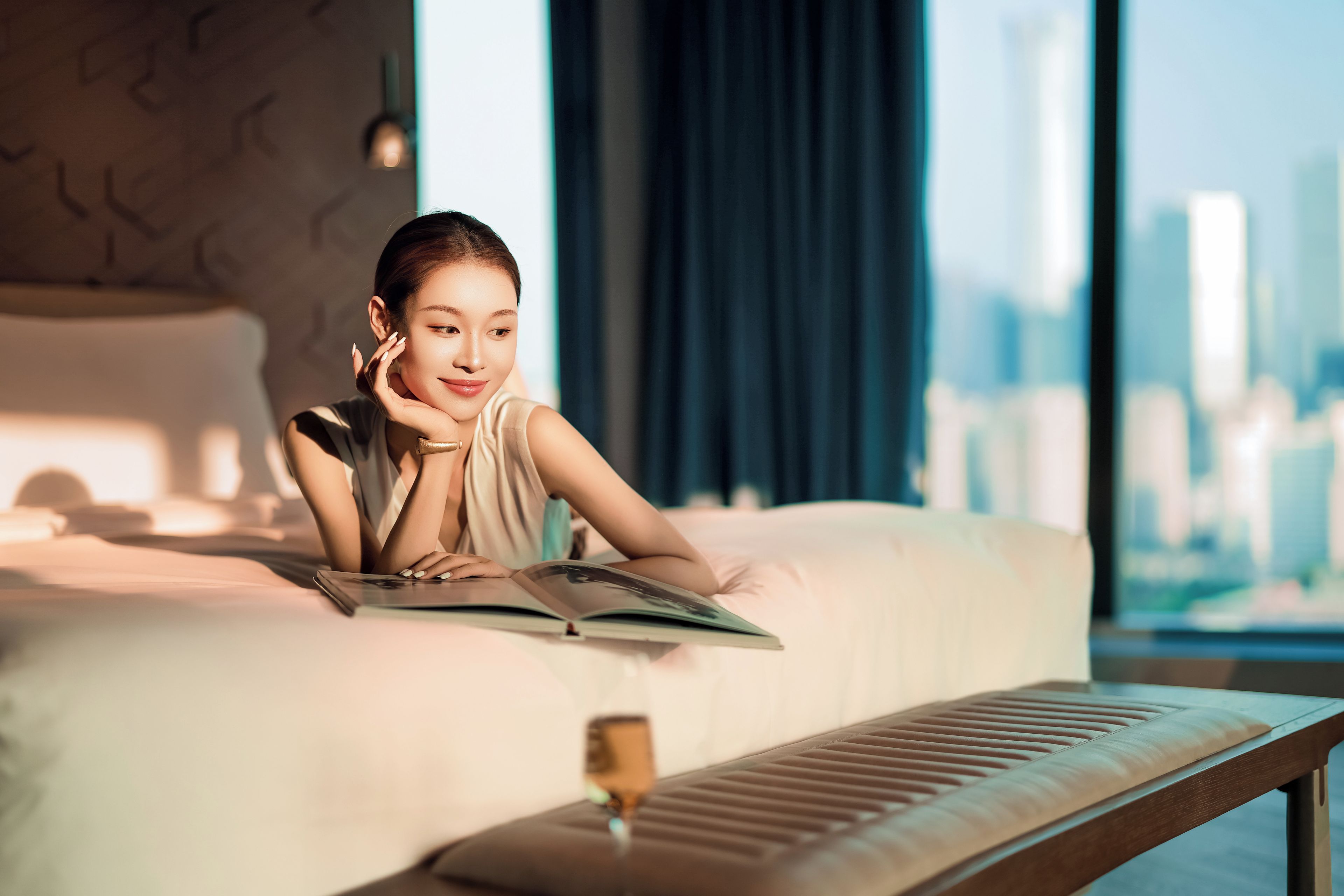 A person lying on a bed with a book, a wine glass, and a cityscape view through a window.