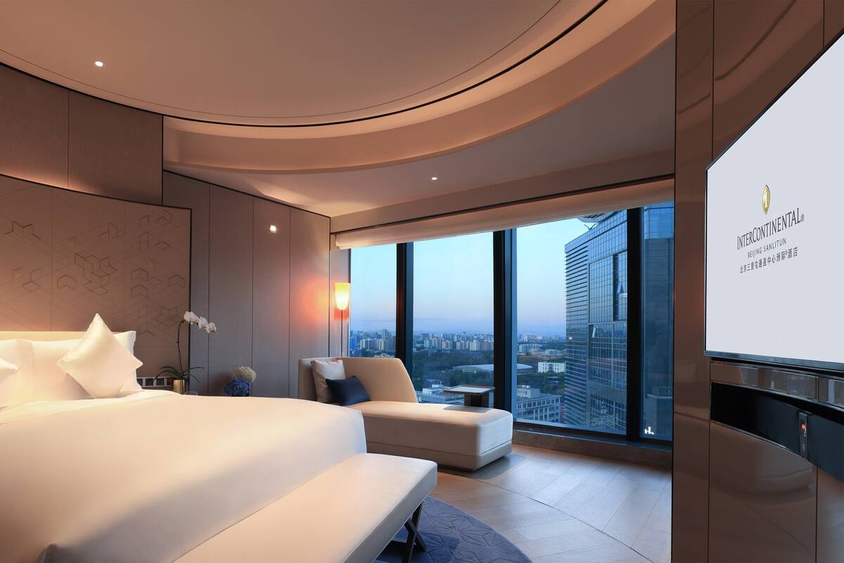 A modern hotel room with a large bed, a seating area, a city view, and a large screen displaying the hotel name.