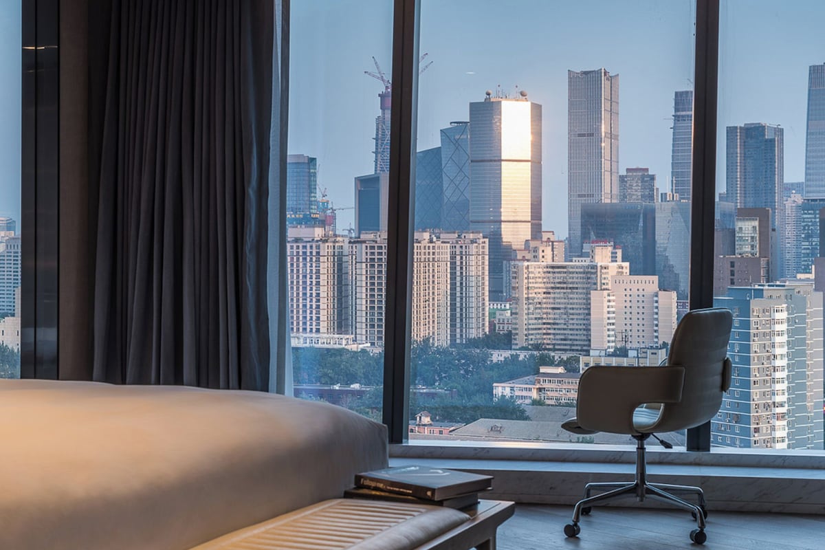 A hotel room with a large window, a bed, a chair, and a view of a cityscape.