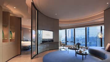 A modern hotel room with a curved ceiling, a large window offering a city view, a flat-screen TV, and a seating area with a sofa and coffee table.