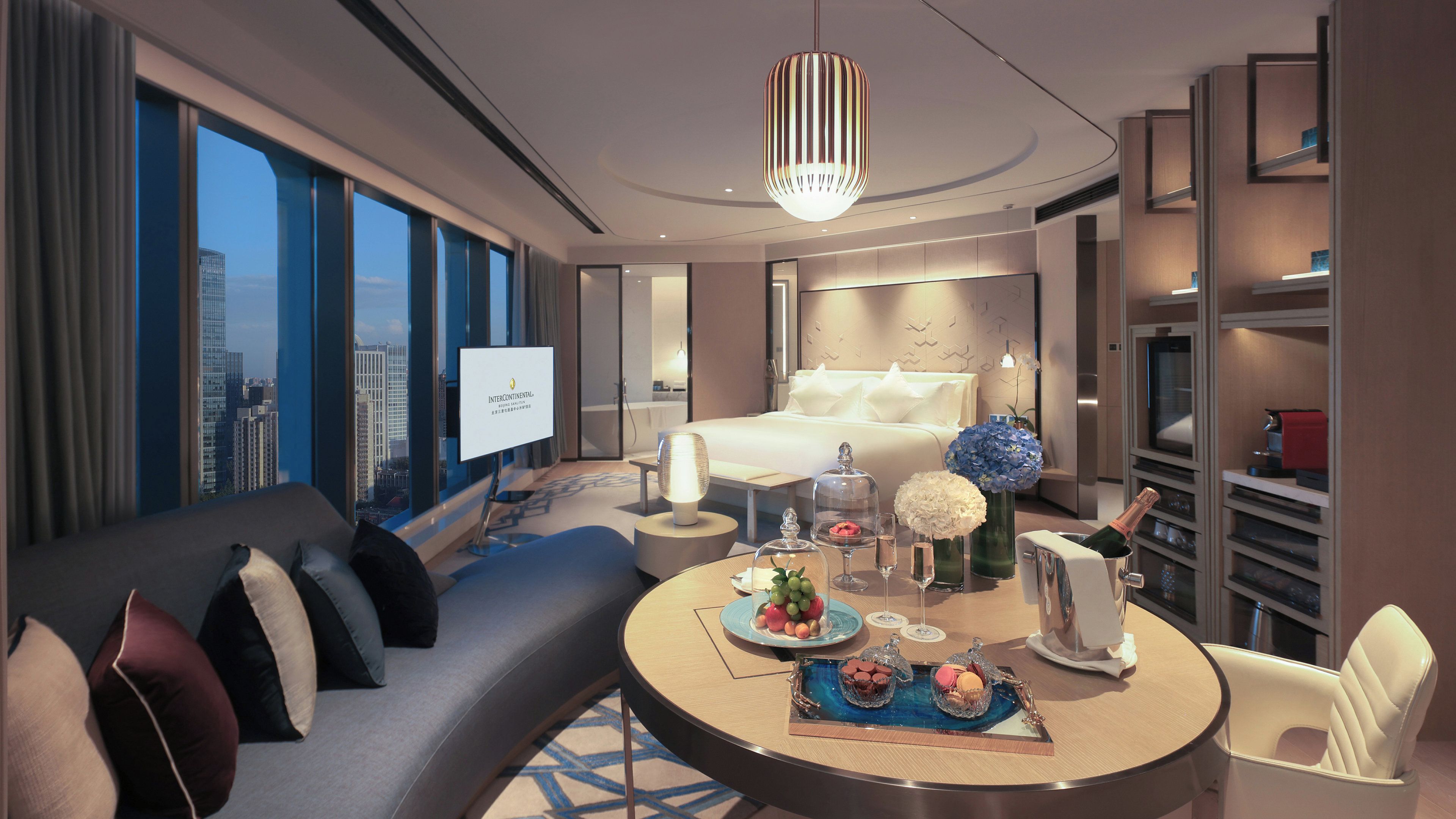 A modern hotel room with a bed, a sofa, a dining table with food and drinks, and a city view through large windows.