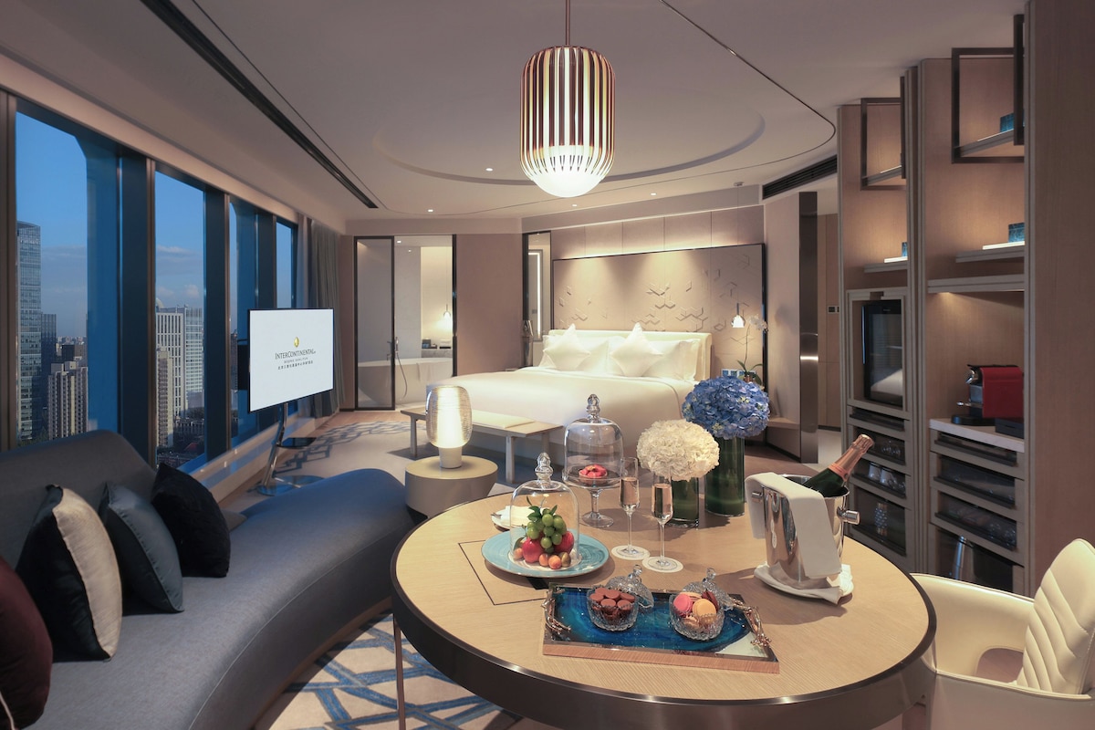 A modern hotel room with a bed, a sofa, a dining table with a cake and flowers, and a city view.