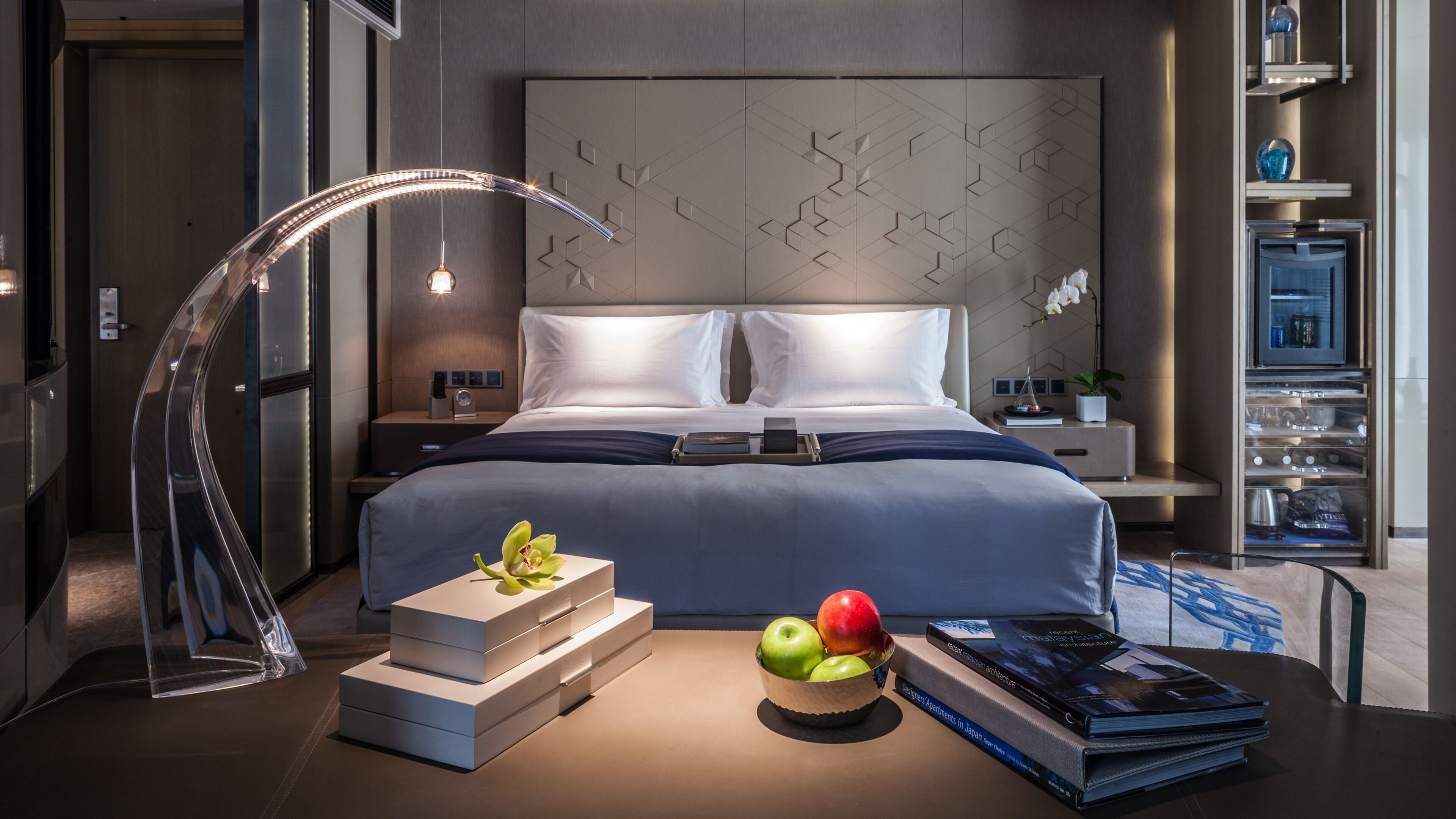 A modern hotel room with a neatly made bed, a bedside table with a lamp, a small plant, and a tray with apples and books.