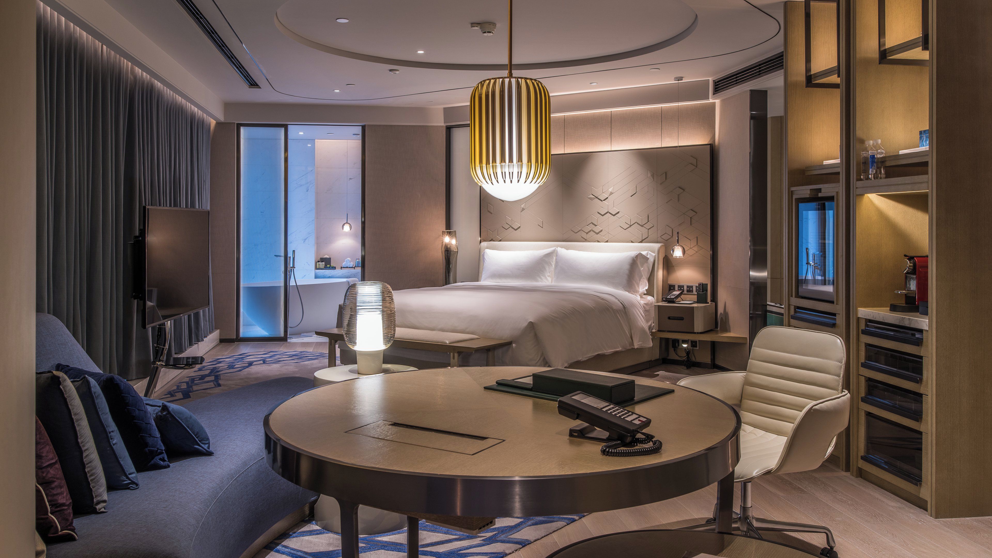 A modern hotel room with a bed, bedside table, chair, round table, and a large window.