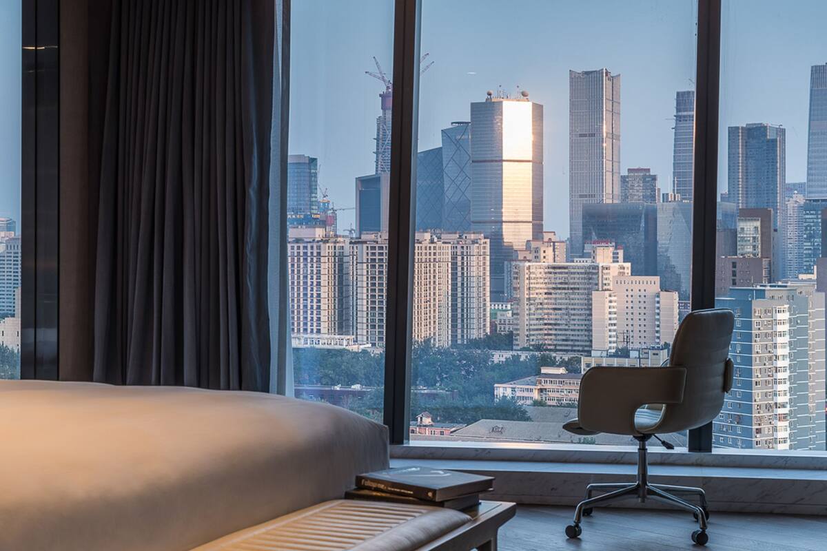 A hotel room with a large window offering a cityscape view, a bed, a chair, and a bedside table.