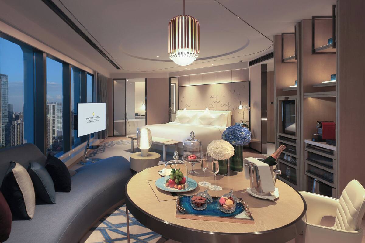 A modern hotel room with a bed, a sofa, a dining table with a cake and flowers, and a city view.