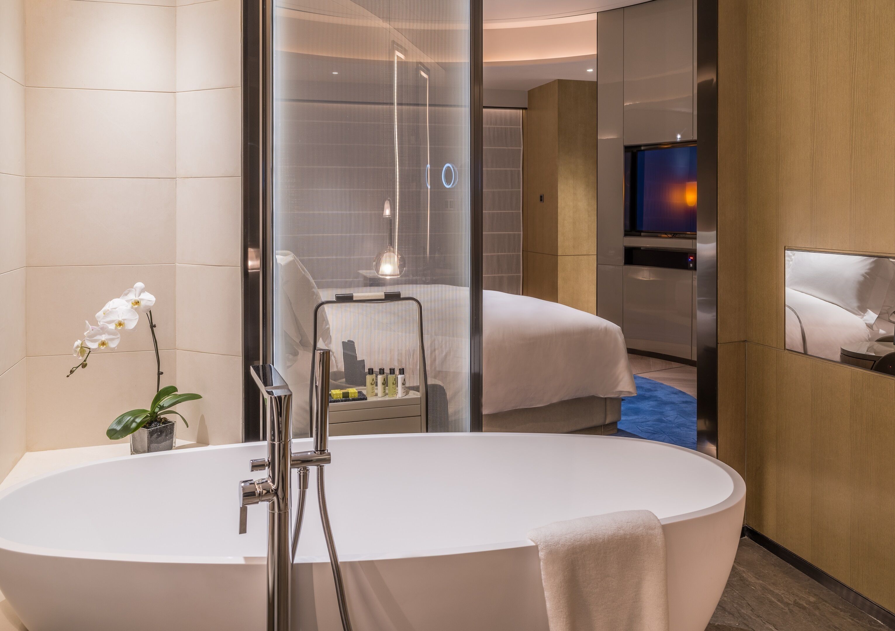 Un baño moderno con una bañera independiente, ducha con mampara de vidrio y un televisor visible en la habitación contigua.