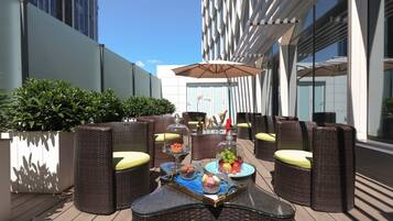 Un espacio al aire libre con mobiliario de mimbre, una mesa con cubierta de vidrio y comida, bebidas, y vistas del edificio de fondo.