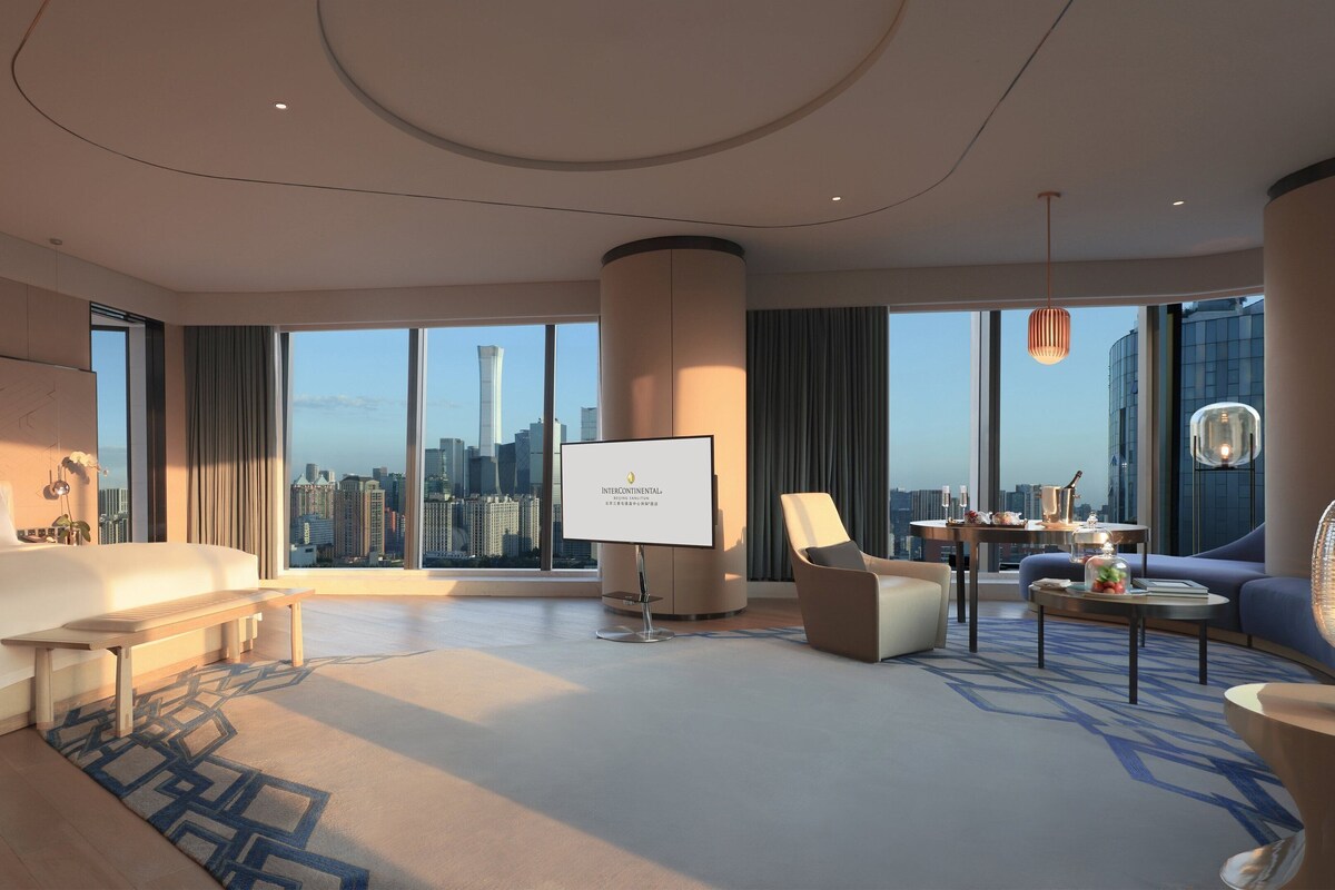 A spacious hotel room with a large bed, a seating area, a TV screen, and a city view.