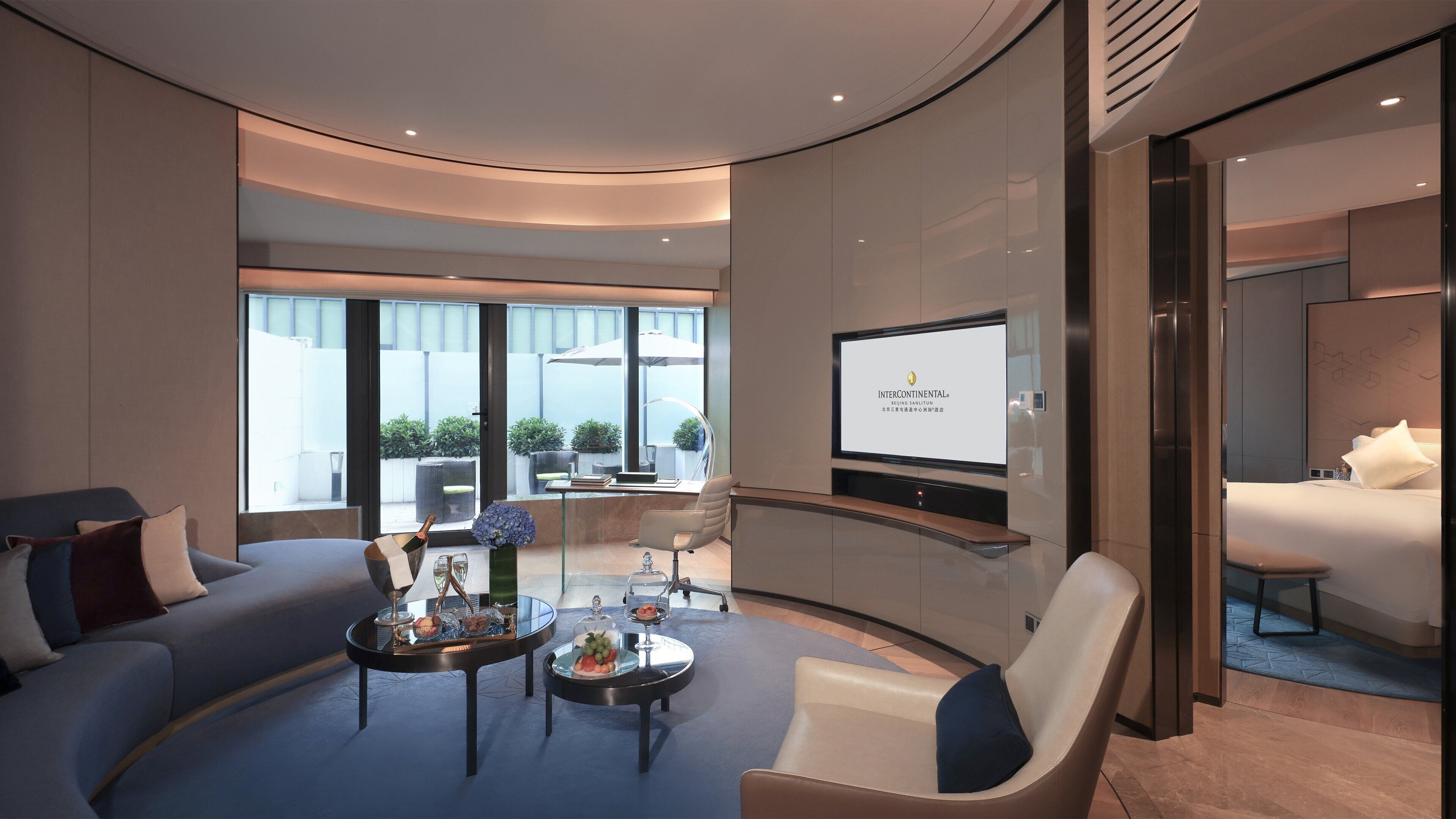 A modern hotel room with a curved layout, a large TV screen, a seating area with a coffee table, and a balcony with a view.