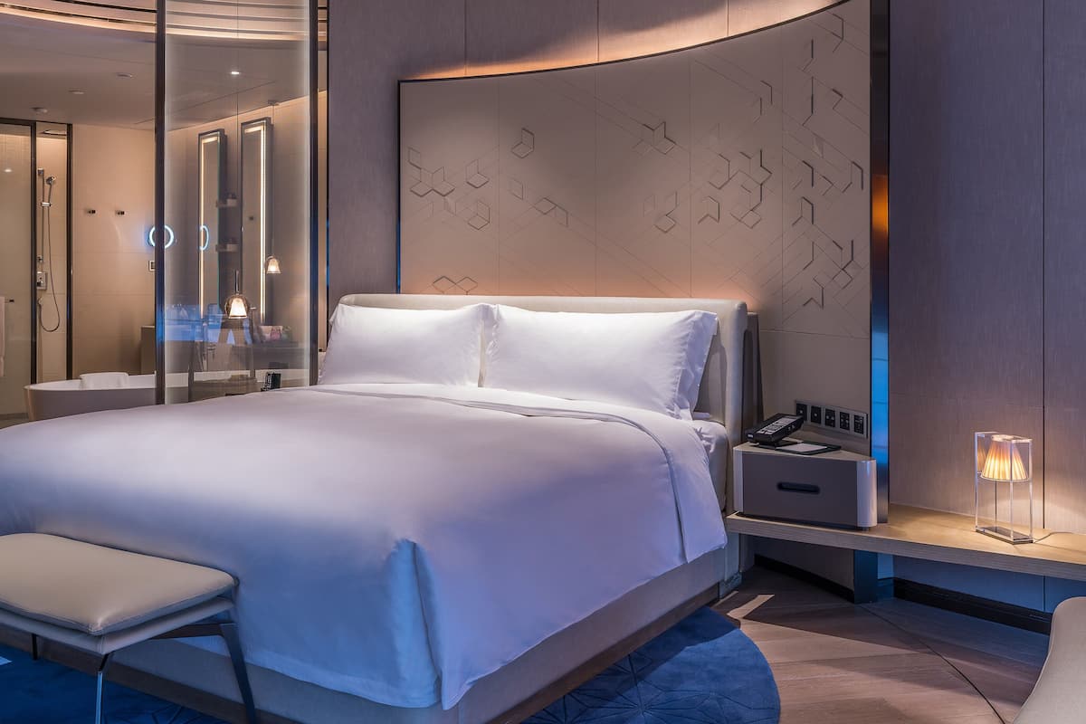 A modern hotel room with a large bed, a bedside table with a lamp, a glass-enclosed shower area, and a built-in wardrobe.