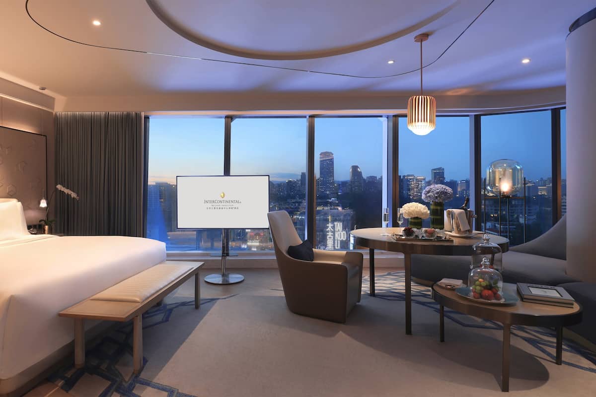 A modern hotel room with a large bed, a desk, and a view of the cityscape.