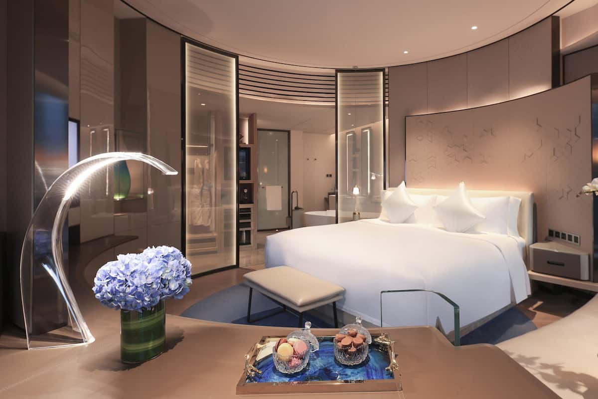 A modern hotel room with a large bed, a vanity with a mirror, a seating area, and a view of the hallway.