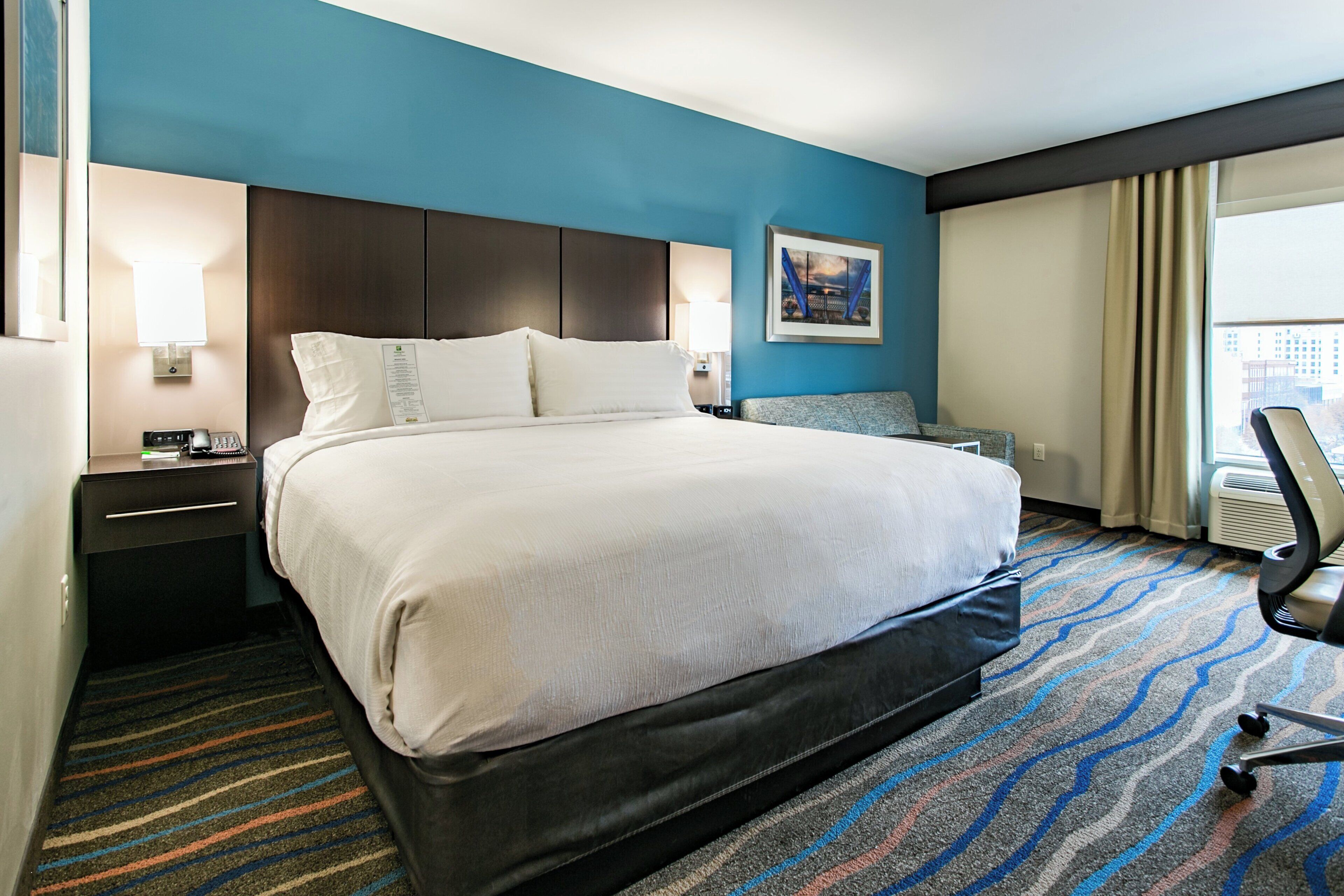 Habitación de hotel con una cama grande, un escritorio con computadora, una silla y una ventana con cortinas.