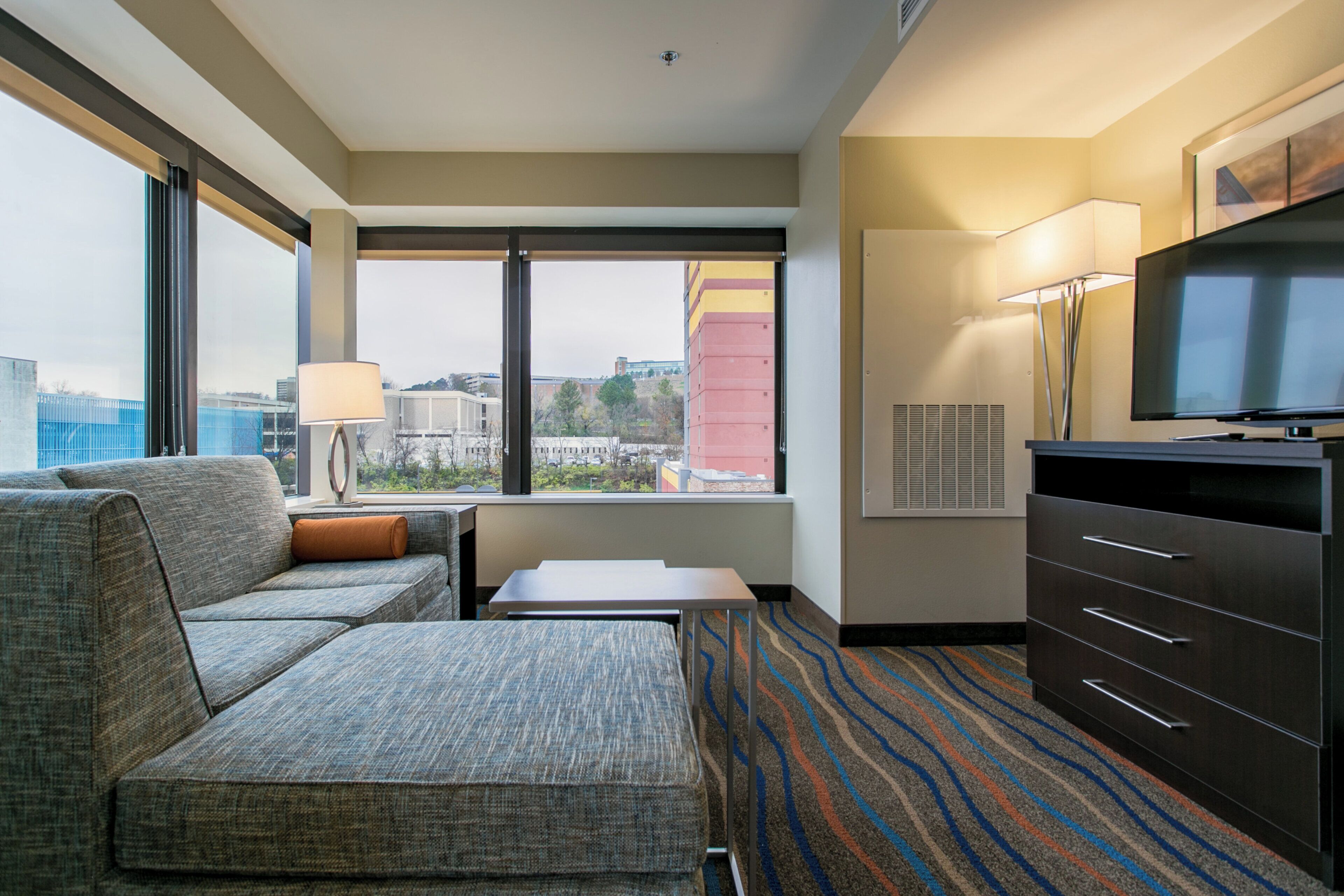 Habitación de hotel con sofá, una mesita, televisor y vistas a la ciudad por la ventana.