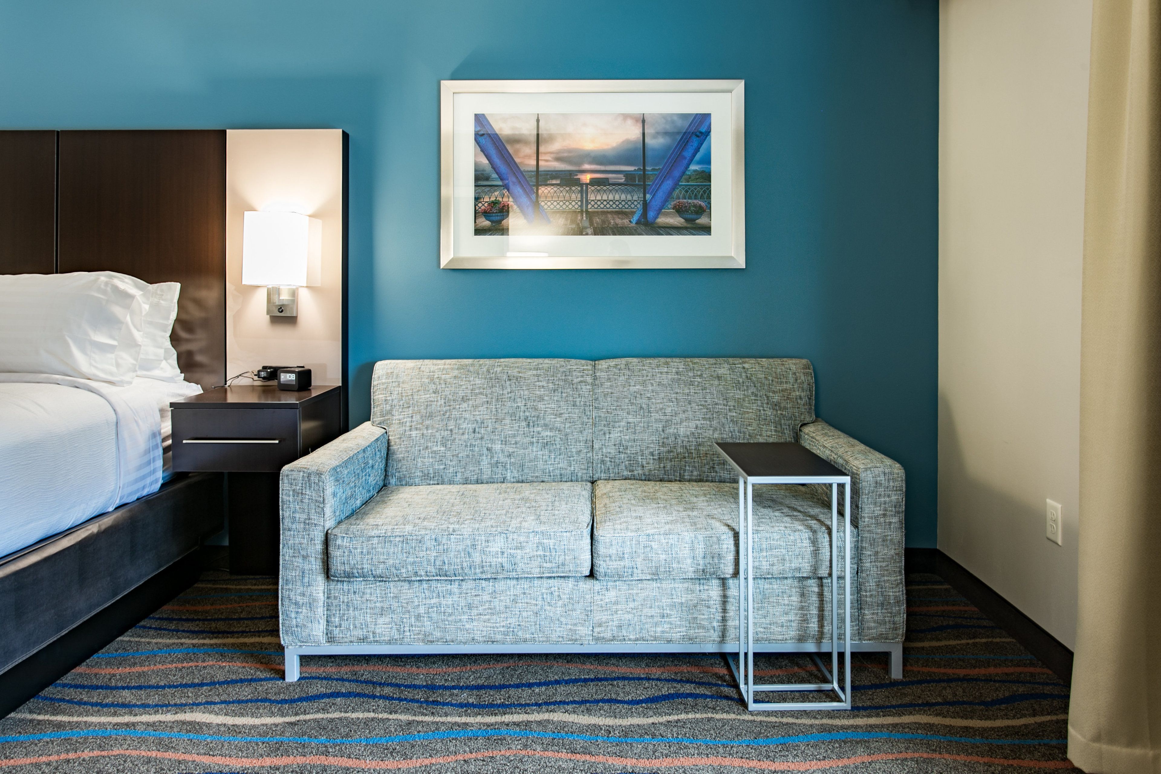 A hotel room with a bed, a sofa, a small table, a lamp, and a framed picture on the wall.
