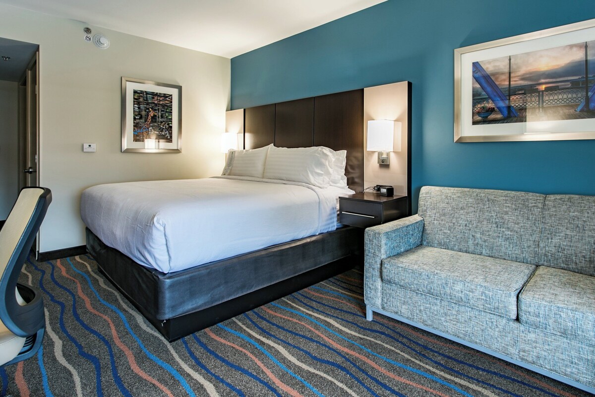 A hotel room with a bed, a nightstand, a chair, and a wall-mounted lamp.