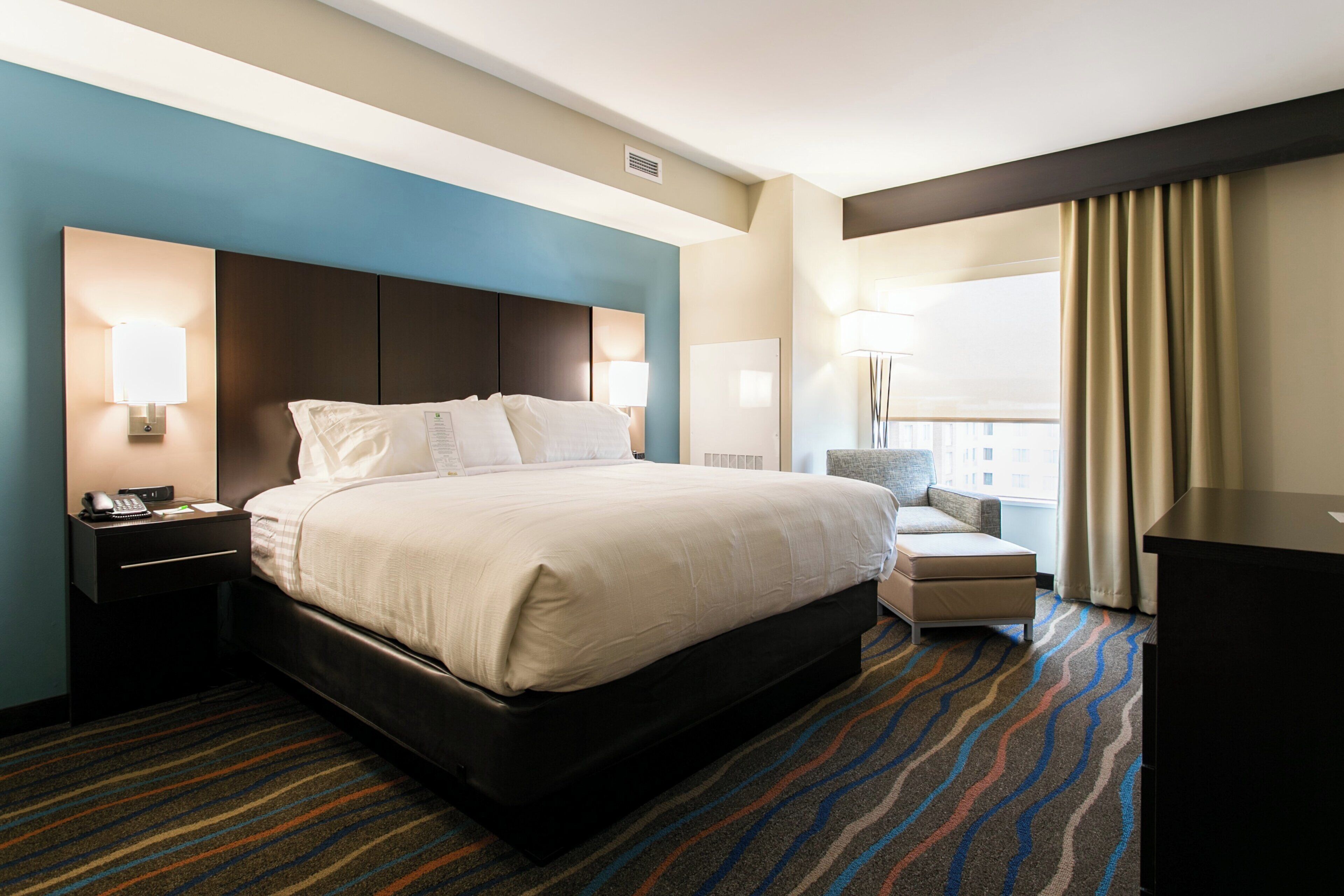 A hotel room with a large bed, a desk with a telephone, a chair, and a desk lamp.