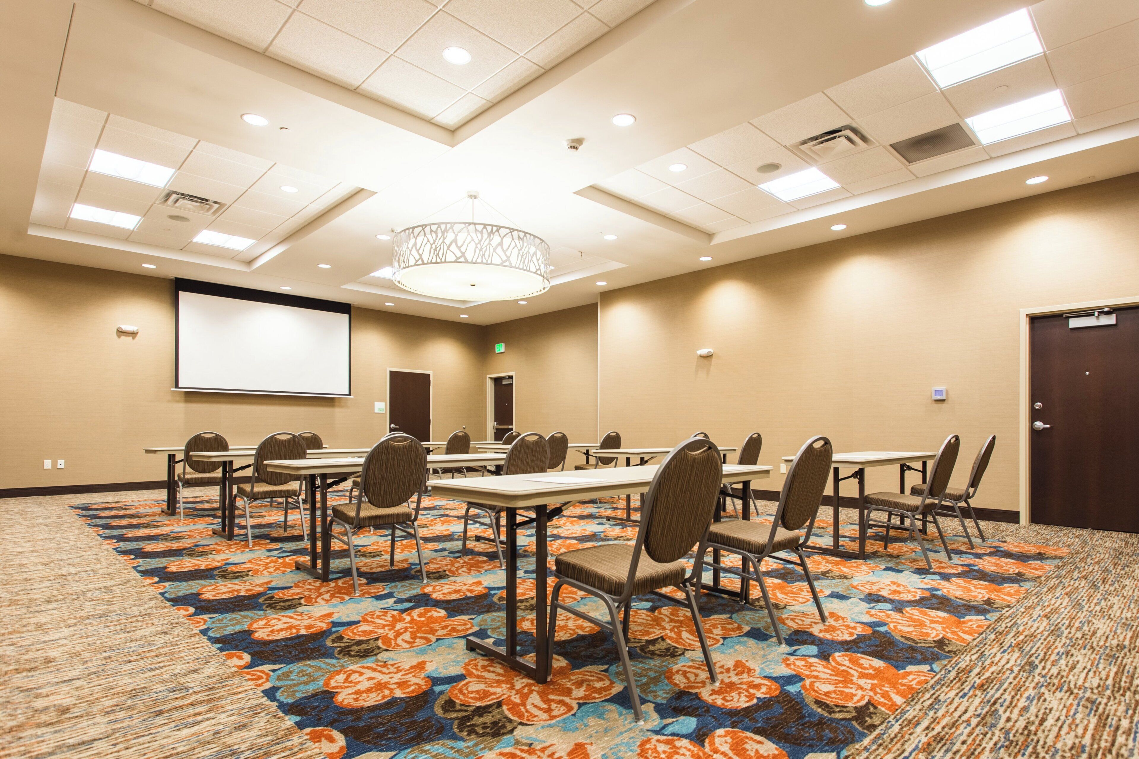 A conference room with a projector screen, tables, chairs, and a carpeted floor.