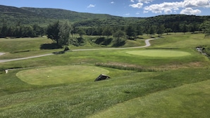 Un parcours de golf avec un chemin sinueux, un green d’entraînement et une colline boisée en arrière-plan.