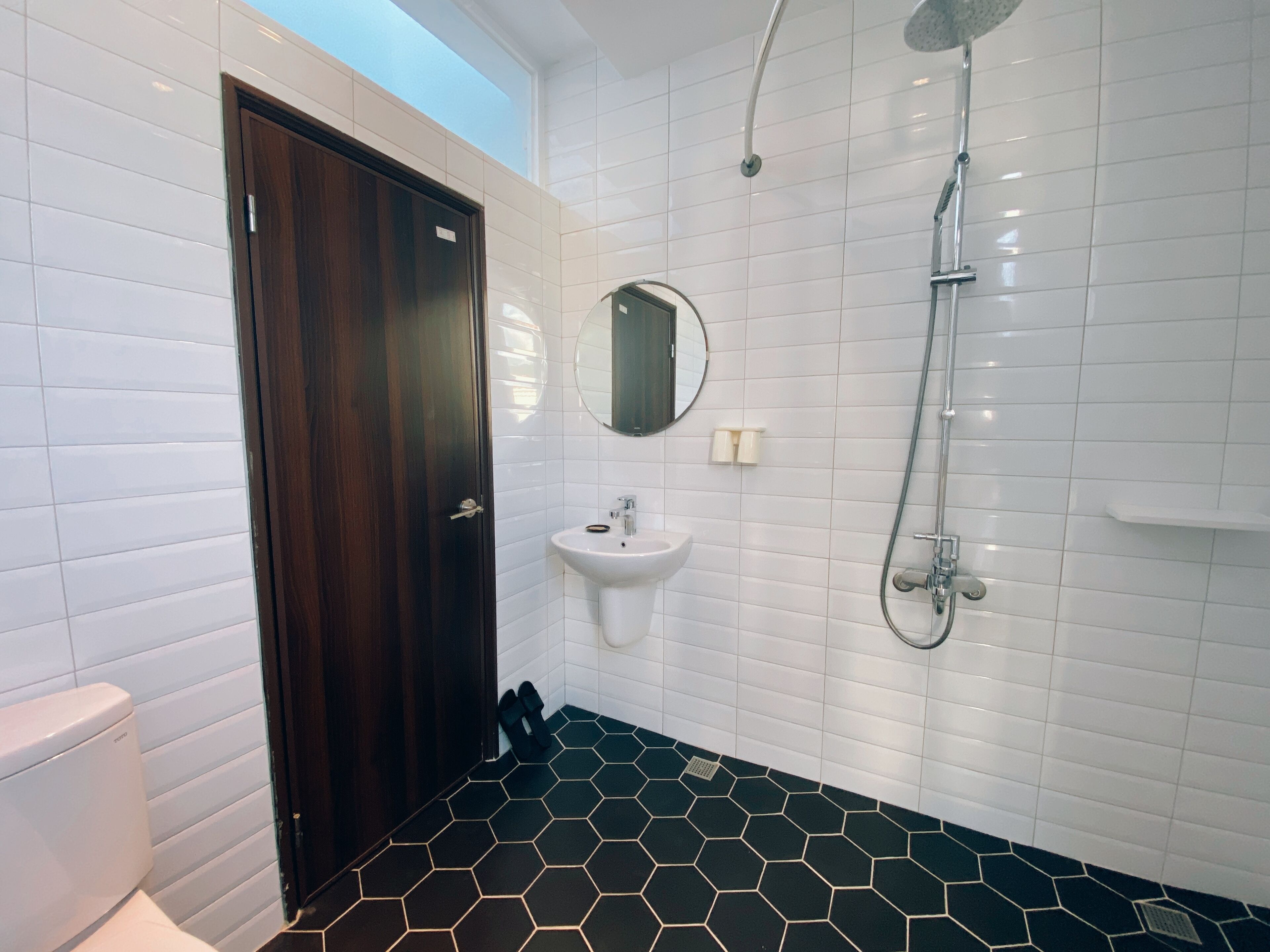 A bathroom with a dark wooden door, white tiled walls, a hexagonal patterned floor, a sink, a mirror, and a shower.