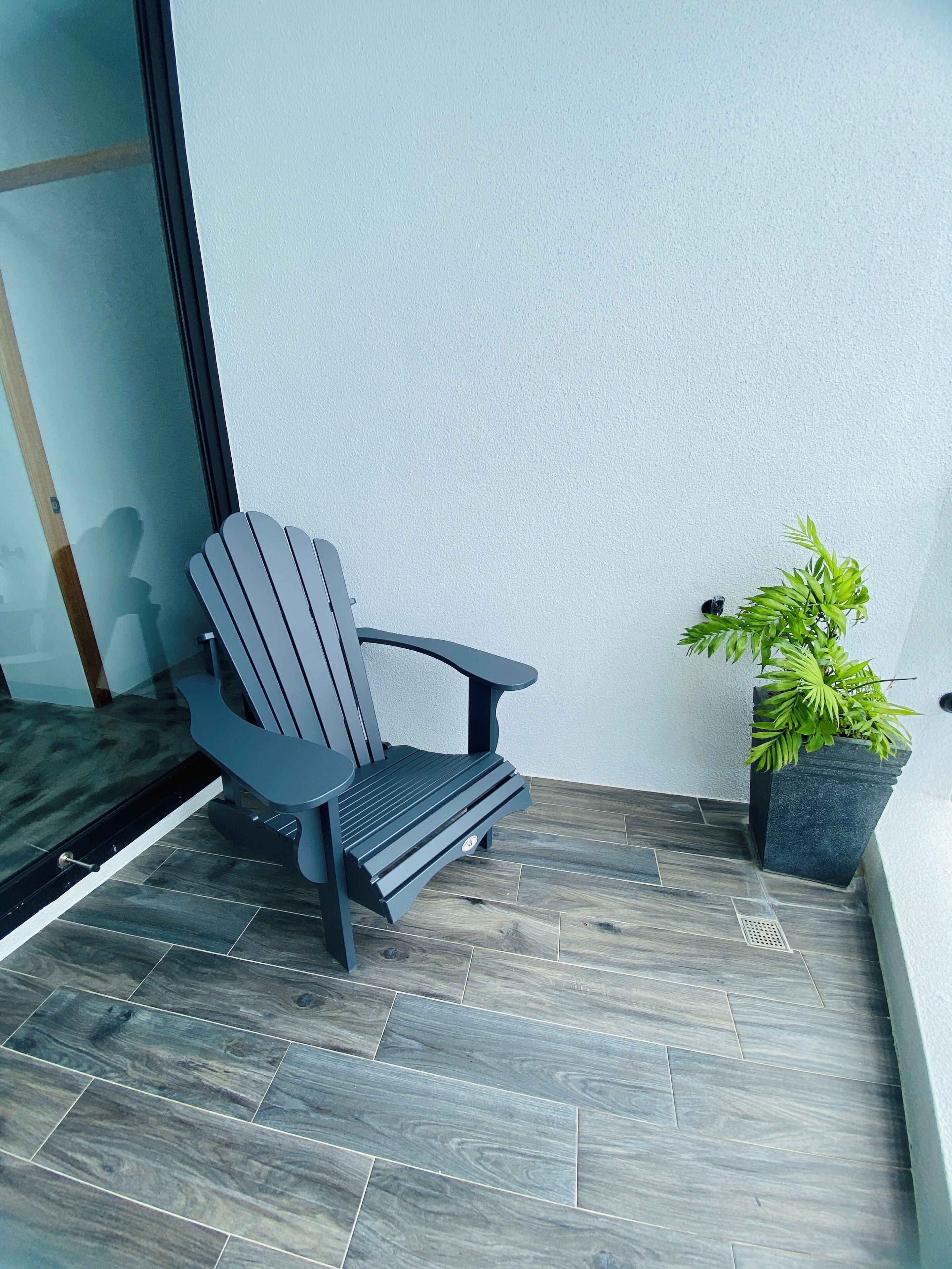 Una silla Adirondack gris en un balcón con piso de baldosas y una planta en maceta al lado.