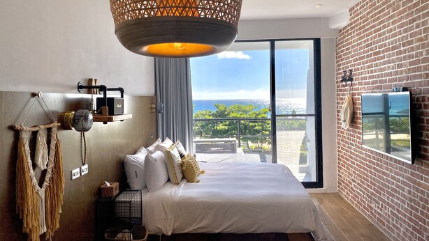 A modern bedroom with a large bed, a hanging light fixture, a macrame wall hanging, and a view of the ocean through a large window. - Minimi Inn - SailRock Beach House (Hengchun)