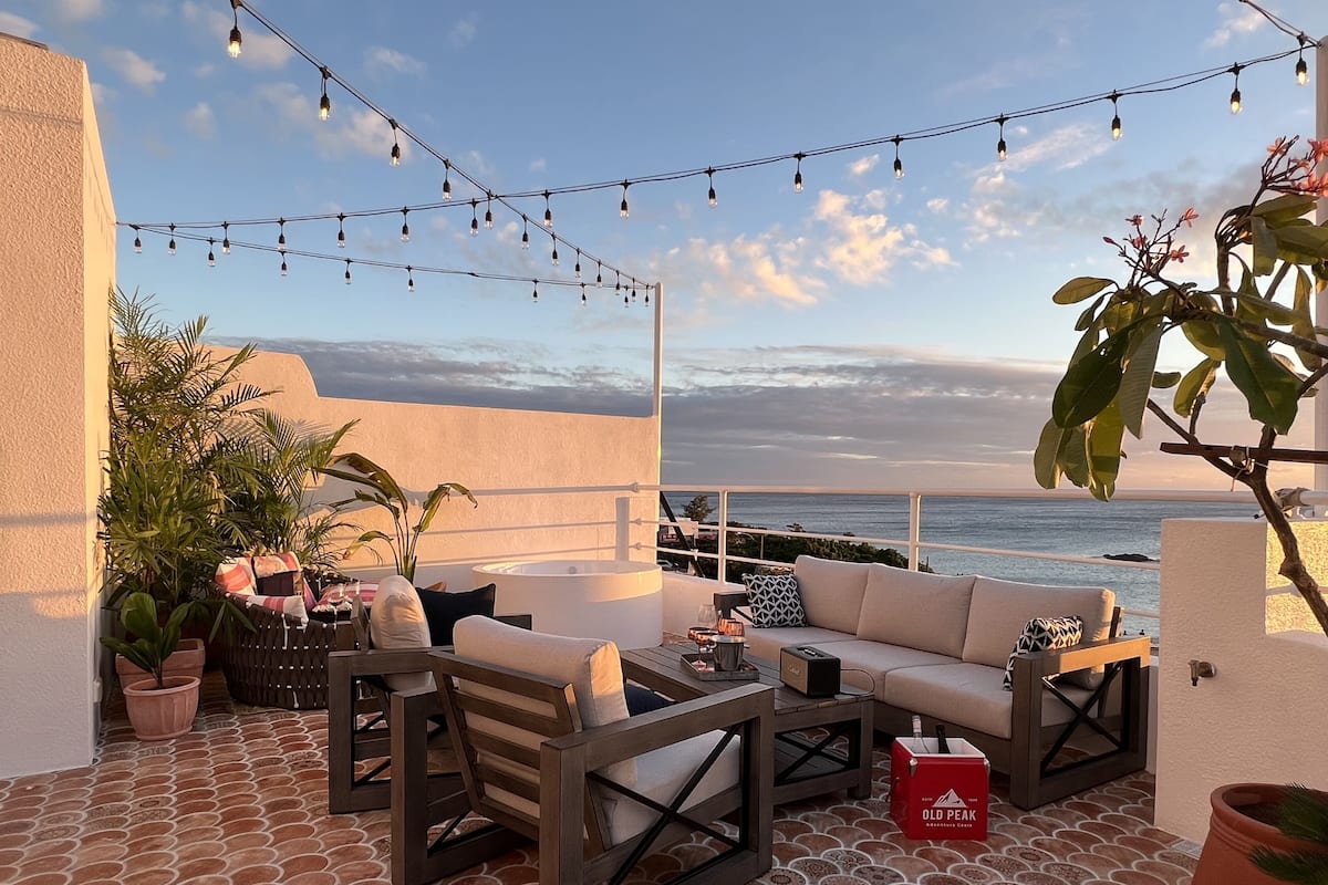 A terrace with outdoor seating, a hot tub, and string lights against a coastal backdrop.