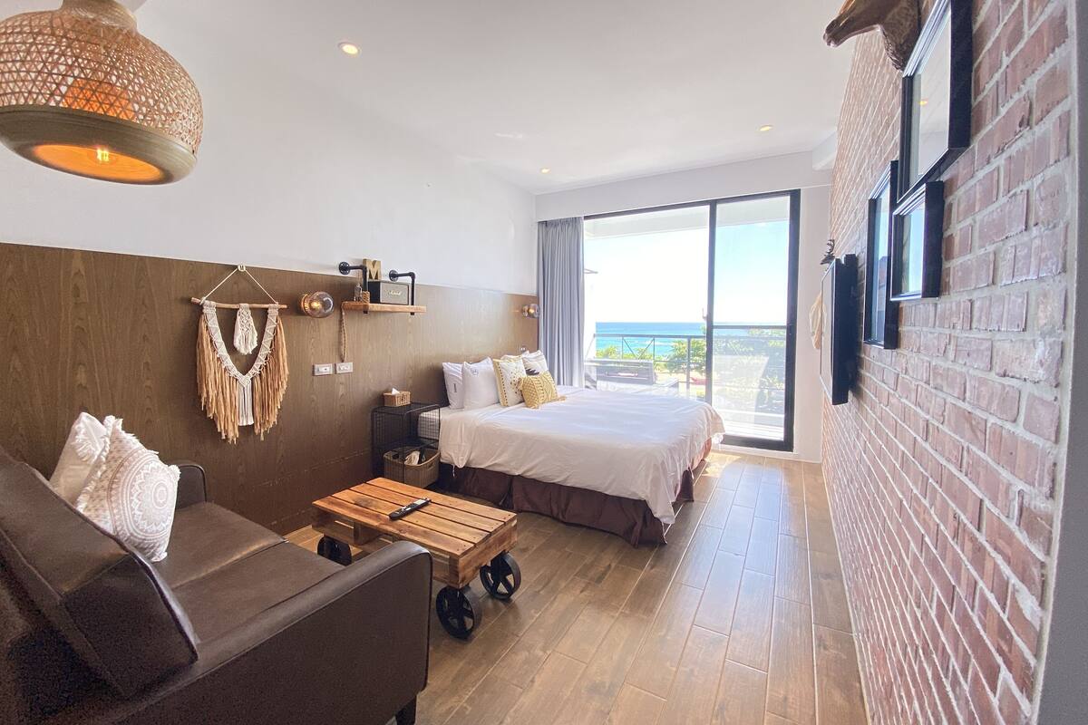 A modern hotel room with a large bed, a sofa, a wooden coffee table, and a view of the ocean.
