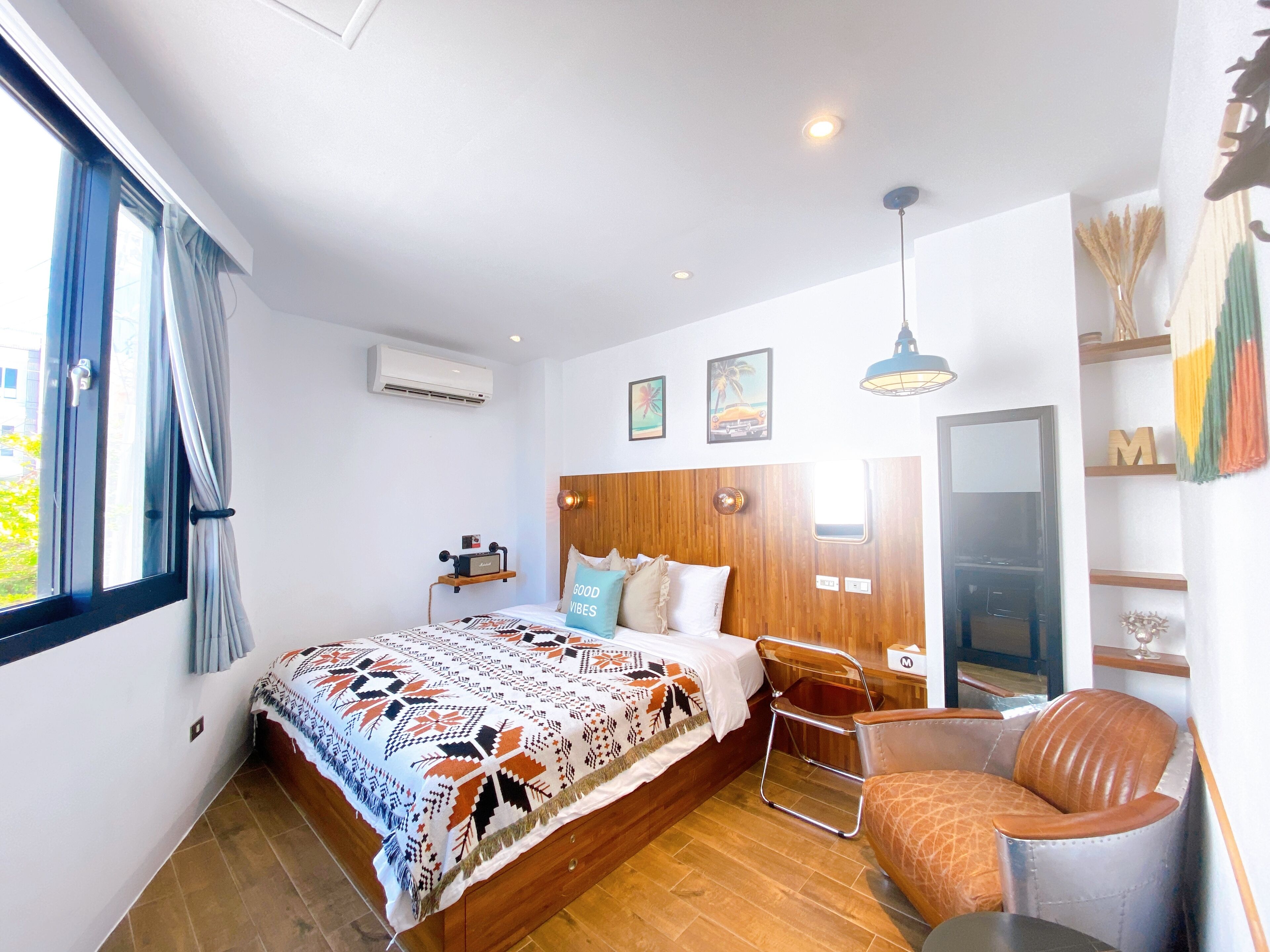 Une chambre à coucher moderne avec une tête de lit en bois, un grand lit avec une literie à motifs, une table de chevet, une chaise, une armoire et une fenêtre avec des rideaux.