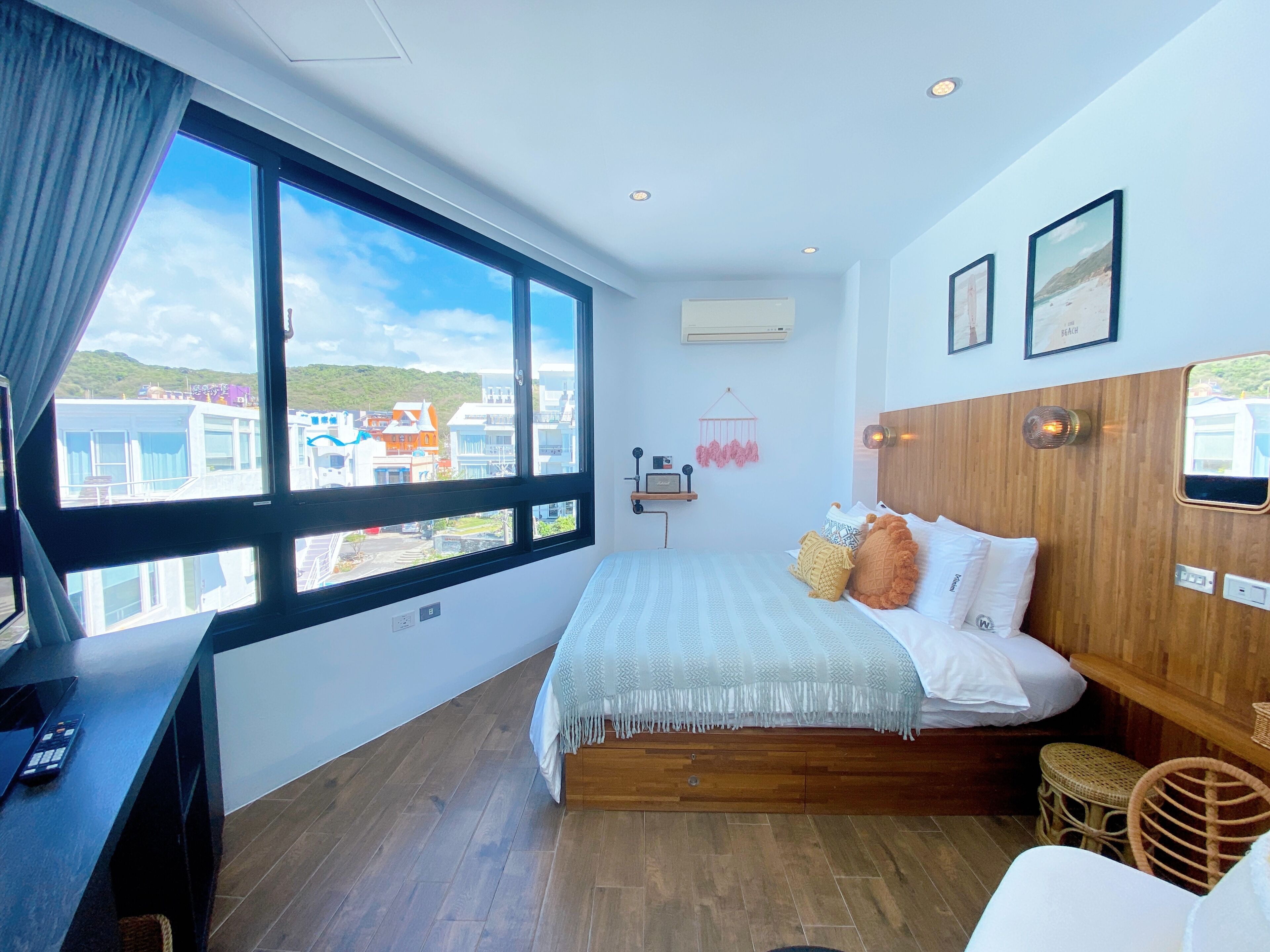 A modern bedroom with a large window, wooden floor, and a bed with white bedding and colorful pillows.