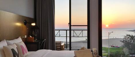 A hotel room with a large bed, a nightstand, a lamp, a wicker basket, and a view of the sunset over the ocean.